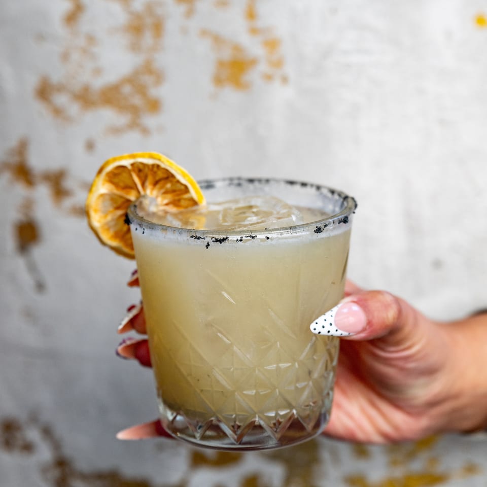 Hand holds a pale citrus cocktail in a textured glass, garnished with a dried orange slice and black-salt rim.