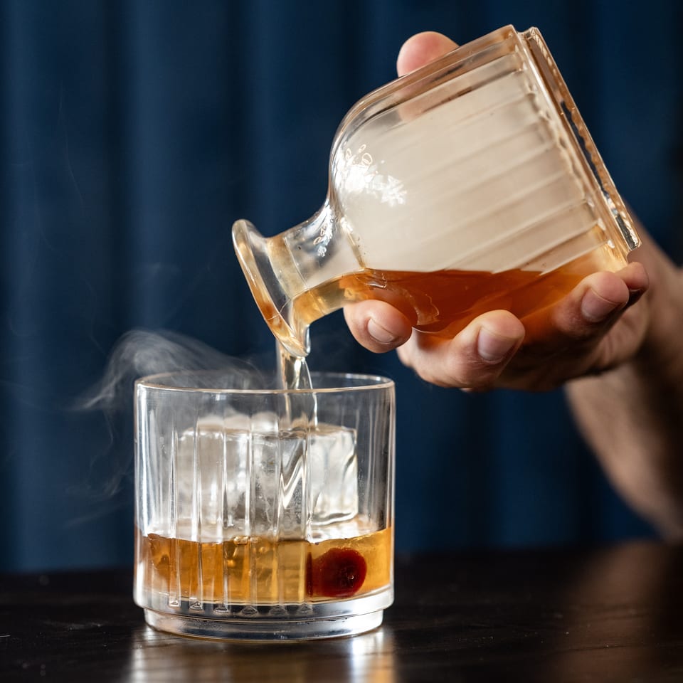 Hands pour an amber cocktail from a glass pitcher into an ice-filled rocks glass garnished with a cherry.