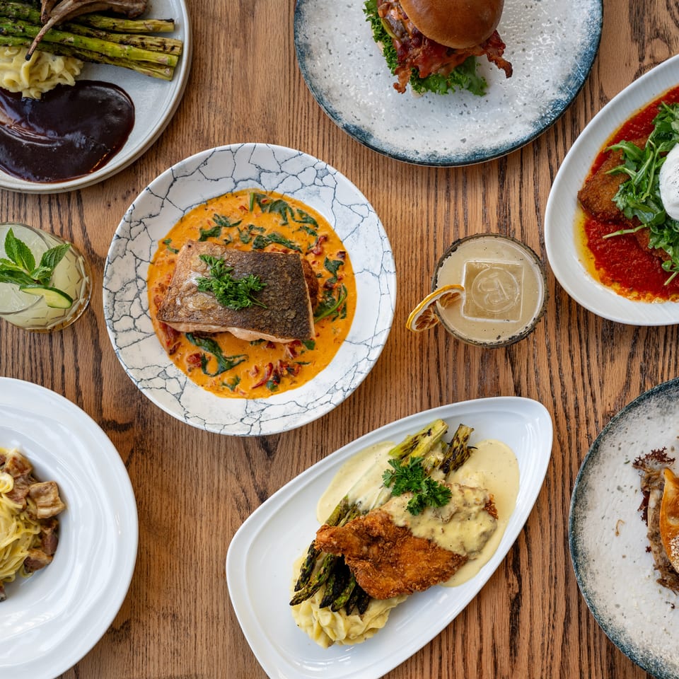 Overhead view of assorted restaurant dishes, including steak, burger, pasta, crispy chicken, fish, burrata, vegetables, and cocktails on a wooden table.
