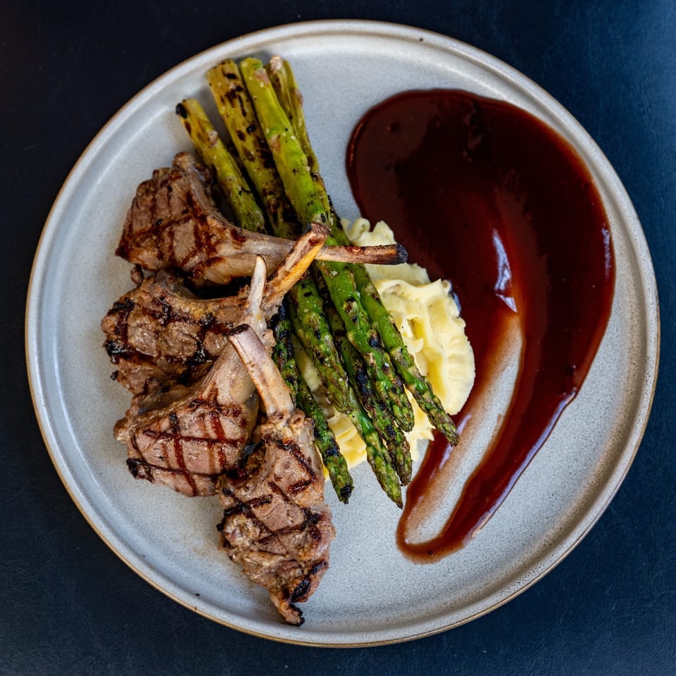 Grilled lamb chops with charred asparagus, mashed potatoes, and red sauce on a ceramic plate.