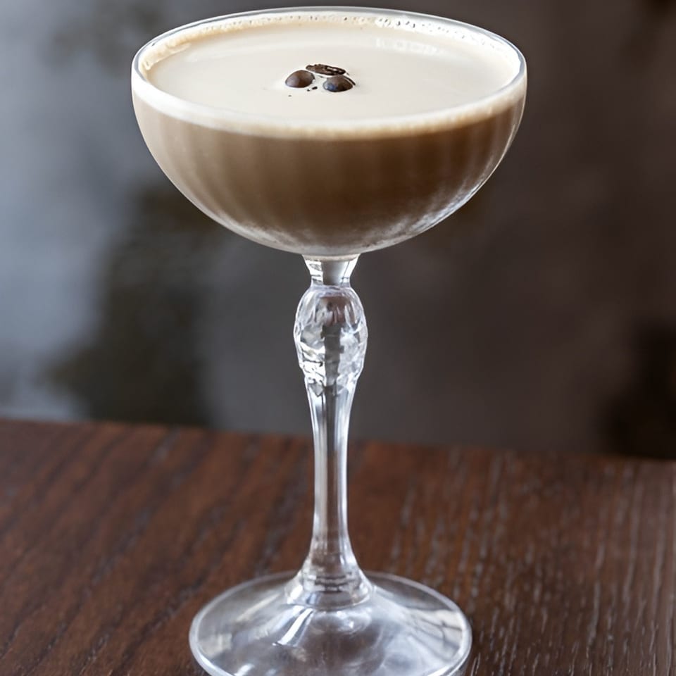 Creamy coffee cocktail in a coupe glass, topped with coffee beans, standing on a dark wooden table.